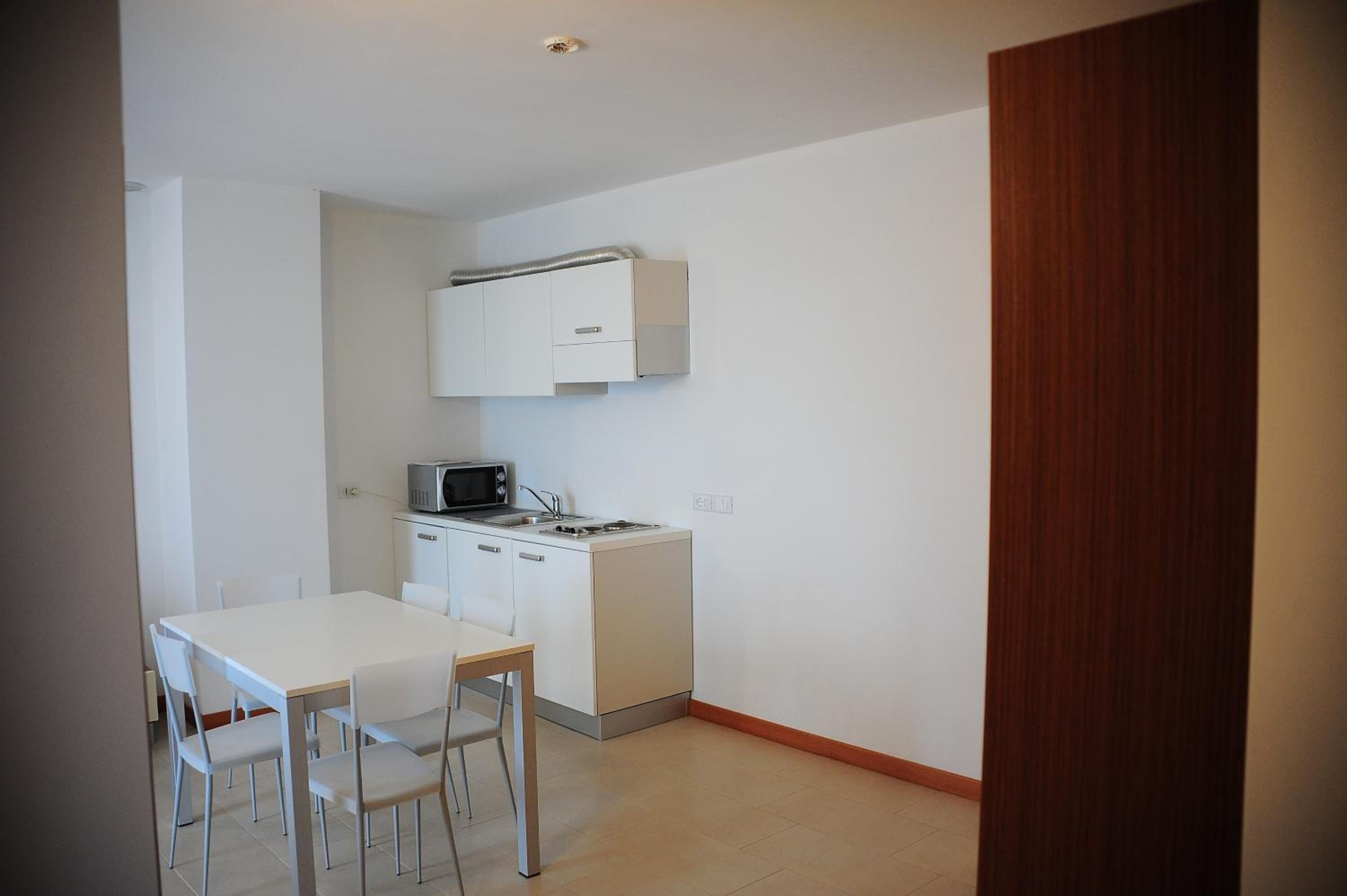Una cocina pequeña y moderna con armarios blancos, un microondas y una estufa. Hay una mesa de comedor blanca con cuatro sillas.