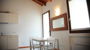 A room with a wooden ceiling, a white table with four chairs, a small kitchenette, and a large window with a mirror above it.