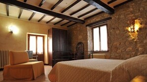 Un dormitorio acogedor con una cama grande, una silla de madera, una pared de piedra y una ventana con luz en cruz.