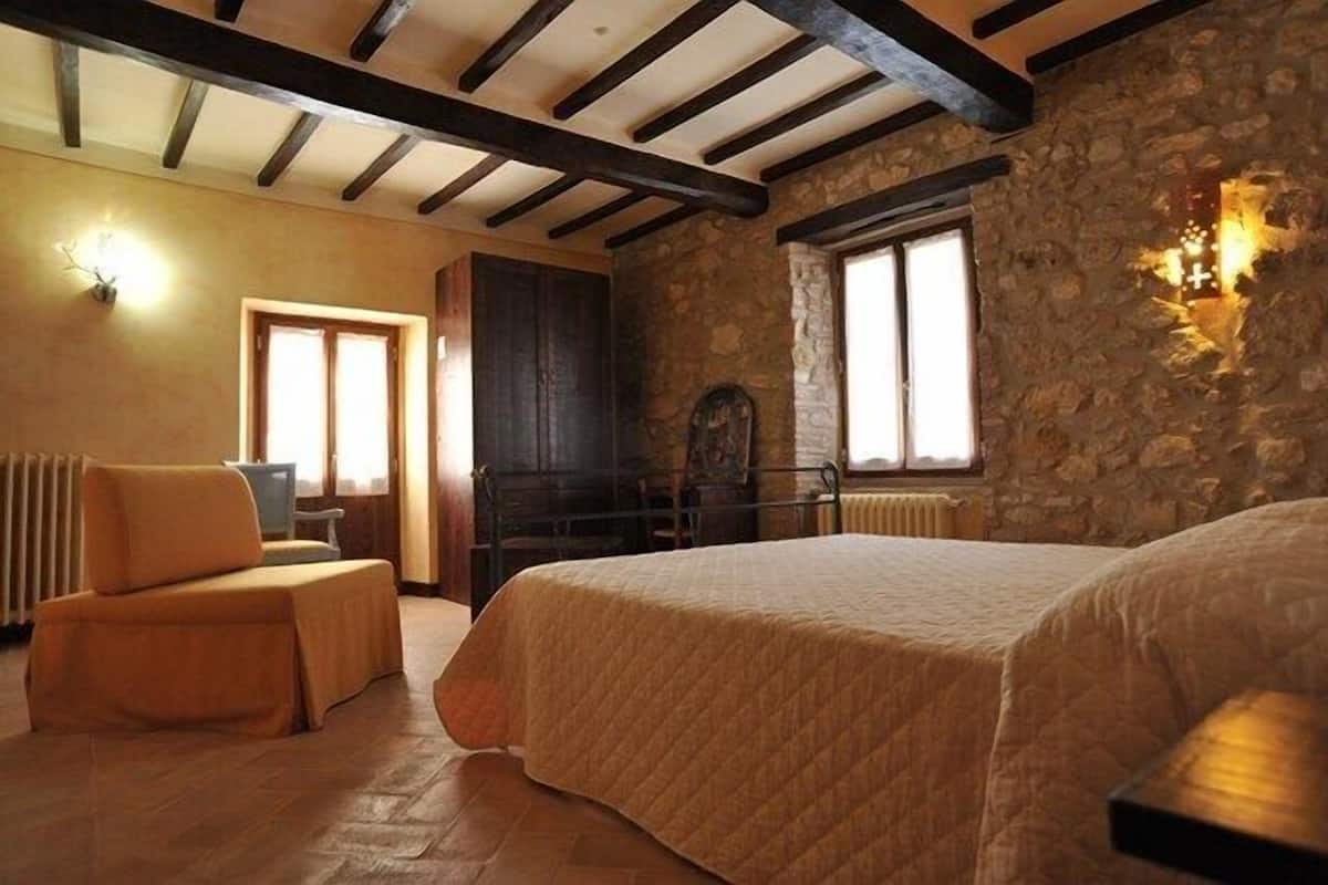 A cozy bedroom with a large bed, a wooden chair, a stone wall, and a window with a cross light.