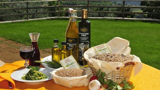 Mesa al aire libre con vistas al paisaje, que incluye una botella de aceite de oliva, una copa de vino tinto, un plato de vegetales verdes y una cesta de cereales.
