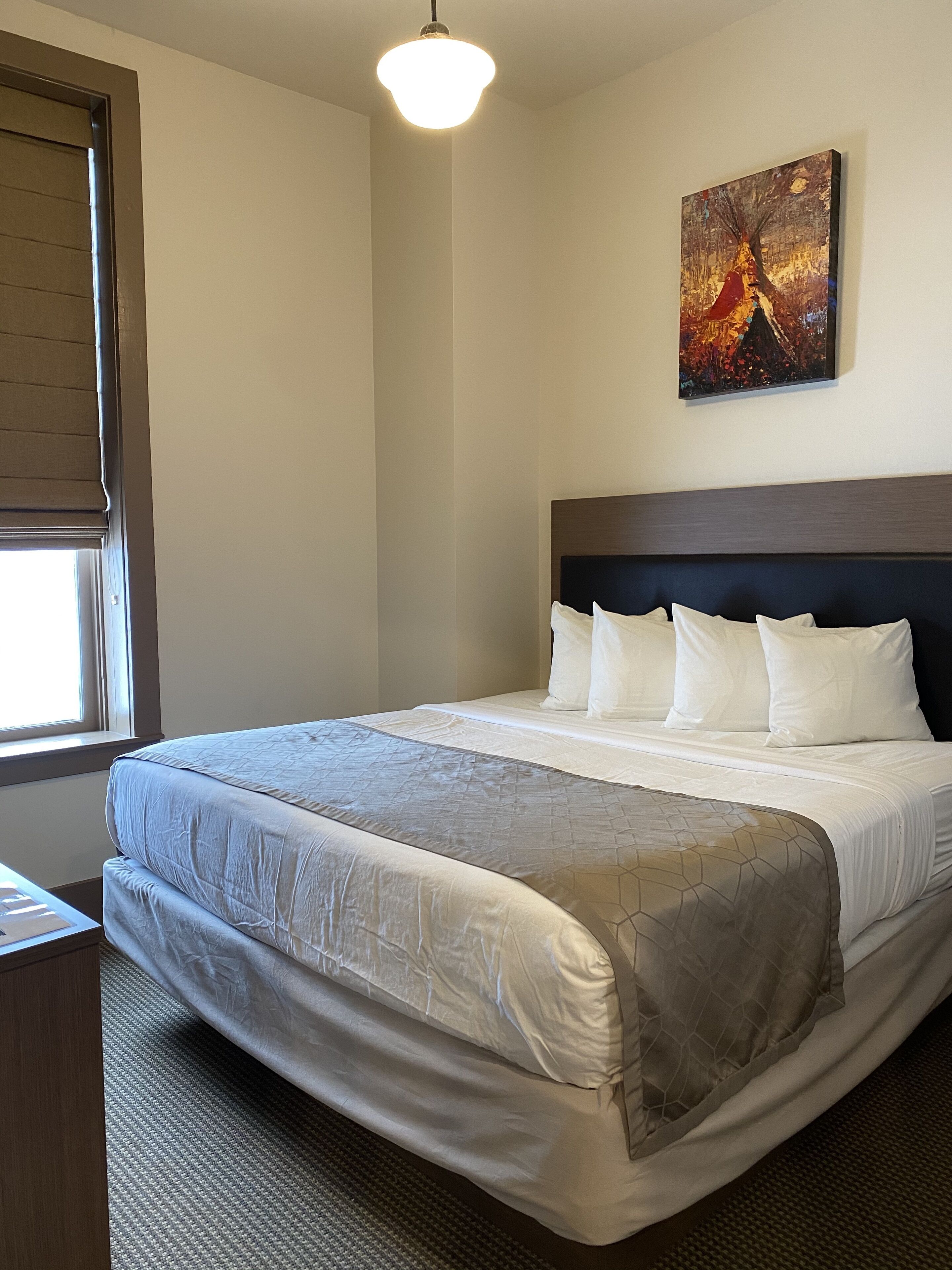 A neatly made bed with a headboard, a painting on the wall, and a window with blinds.