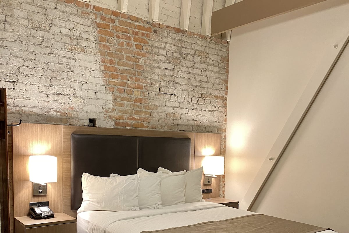 A hotel room with a brick wall, a bed with white bedding, two bedside lamps, a wooden nightstand with a phone, and a red skirted bed.