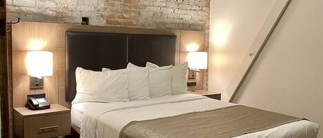 A hotel room with a brick wall, a bed with white bedding, two bedside lamps, a wooden nightstand with a phone, and a red skirted bed.