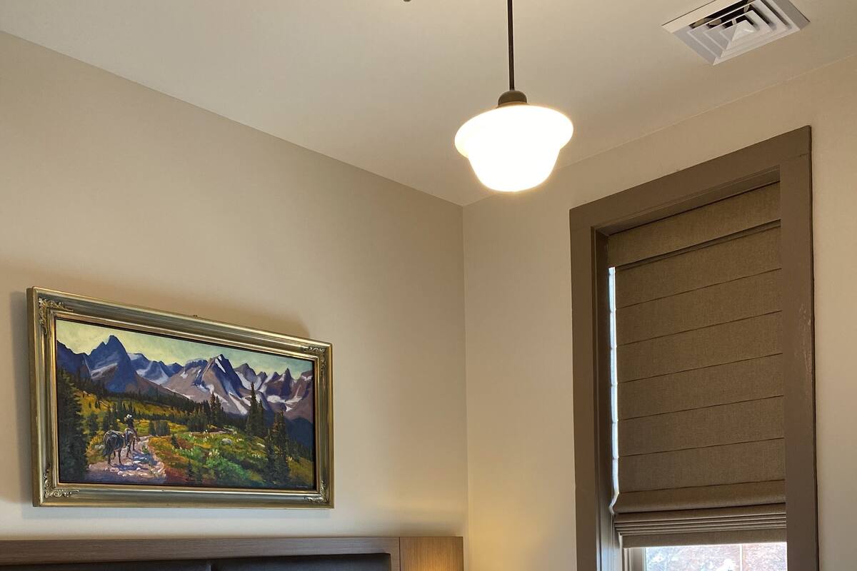 A hotel room with a large bed, a painting of mountains, a nightstand with a lamp, and a window with blinds.