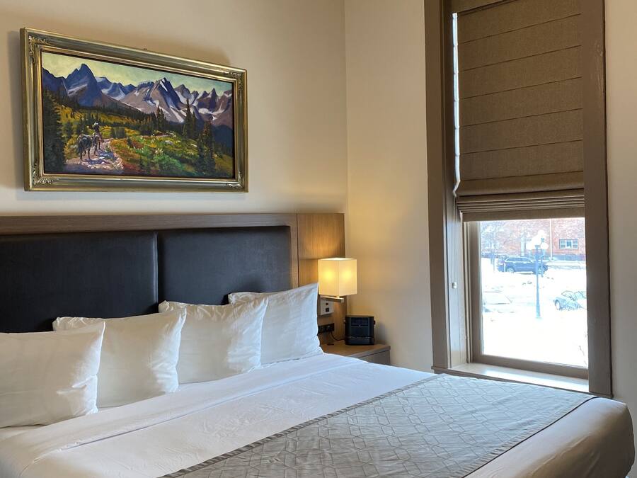 Ein Hotelzimmer mit einem großen Bett, einem Bergpanorama, einem Nachttisch mit Lampe und einem Fenster mit Jalousien.