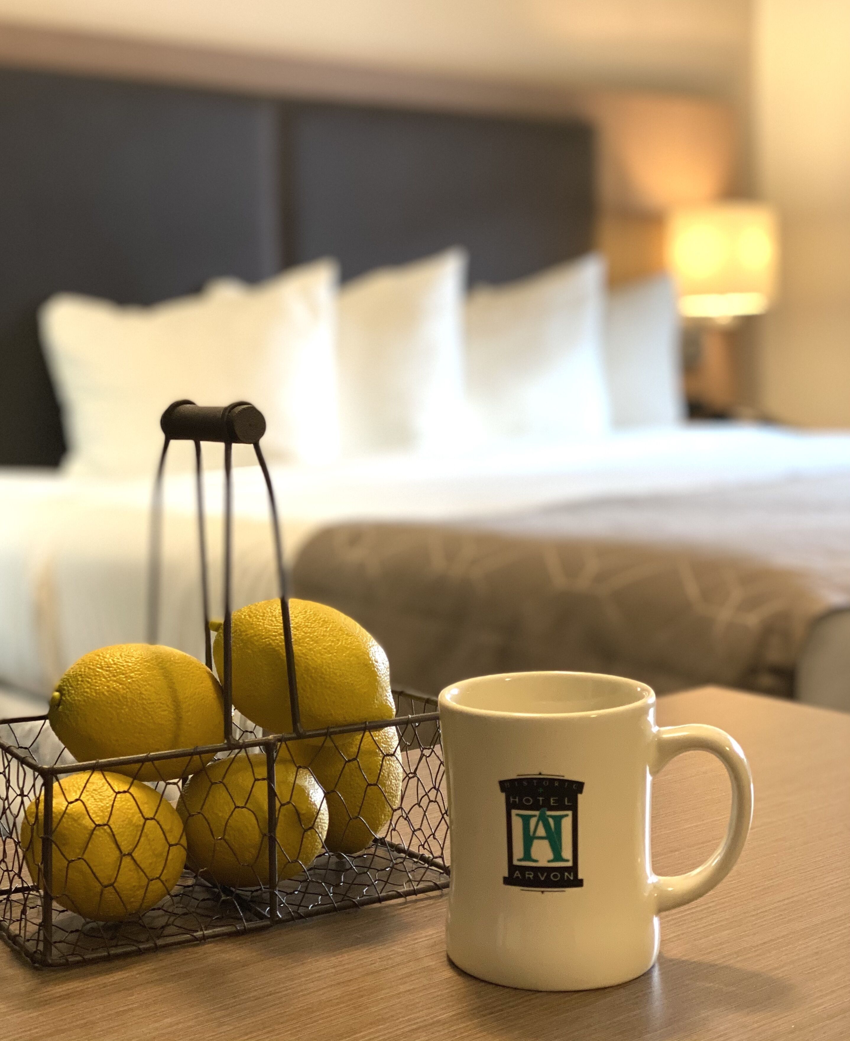 Ett hotellrum med en säng, ett sängbord, en lampa och en korg av metalltråd som innehåller citroner.