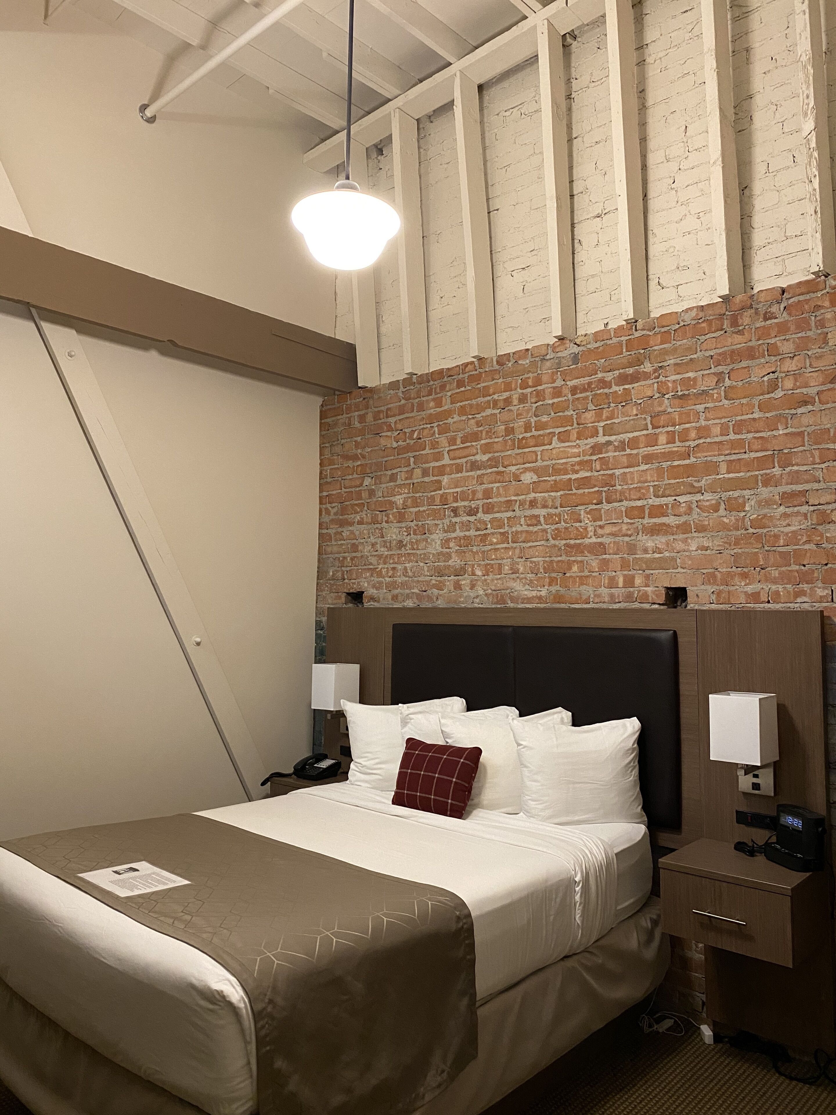 A hotel room with a brick wall, a bed with white linens, a red pillow, a bedside table with a lamp, and a nightstand with a phone and a digital clock.