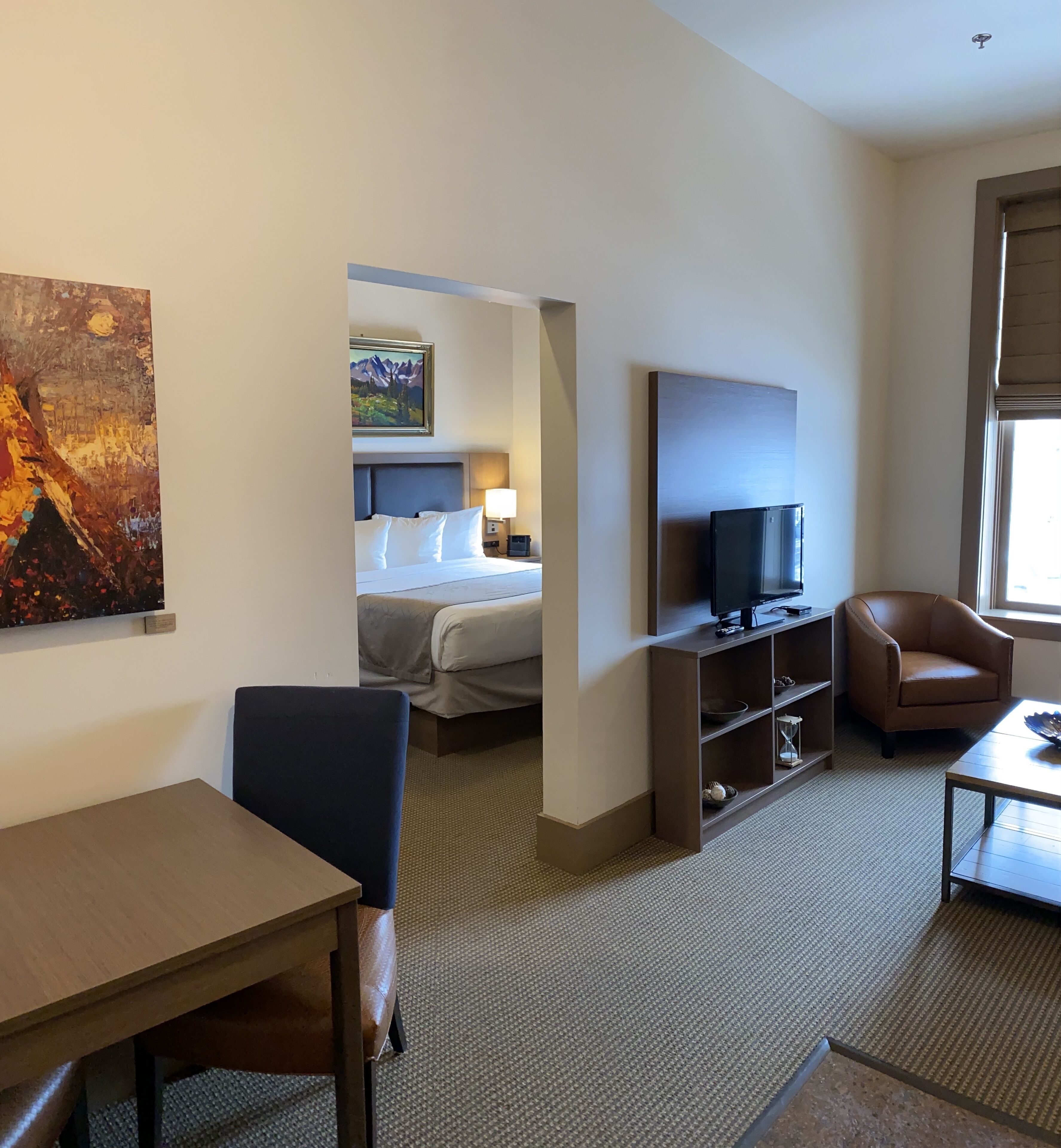 A hotel room with a bed, a TV, a desk, a chair, and a painting on the wall.