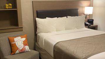 A hotel room with a bed, a nightstand with a lamp and phone, a chair with a pillow, and a shelf with fruits.