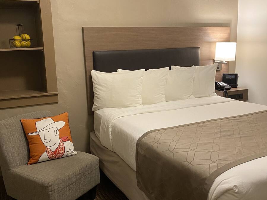 A hotel room with a bed, a nightstand with a lamp and phone, a chair with a pillow, and a shelf with fruits.