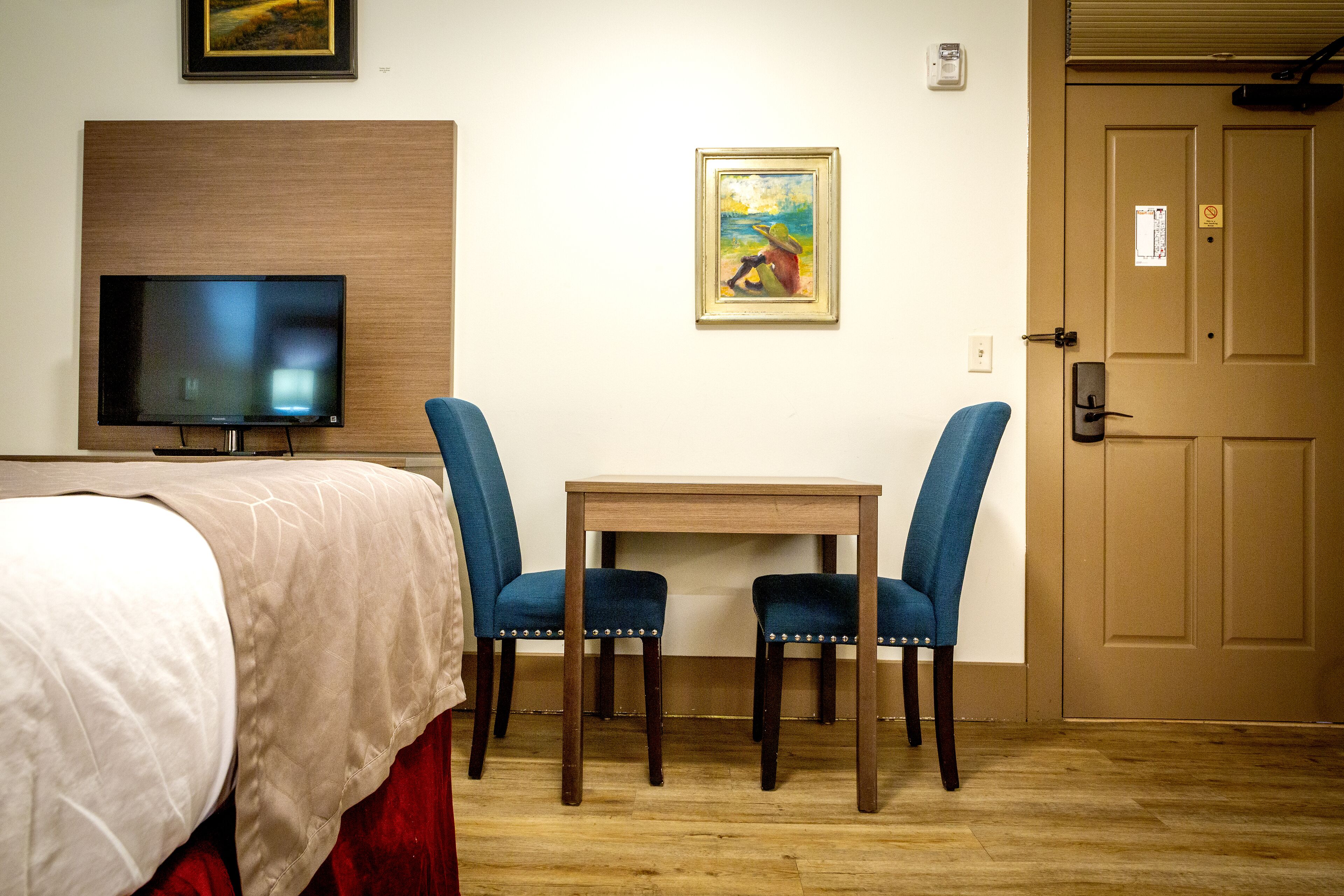 A hotel room with a bed, a television, two blue chairs, a small table, and a door.