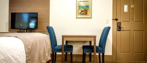A hotel room with a bed, a television, two blue chairs, a small table, and a door.