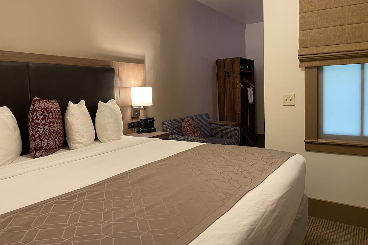 A hotel room with a large bed, a nightstand with a lamp, a seating area with a chair and a small table, and a wardrobe.