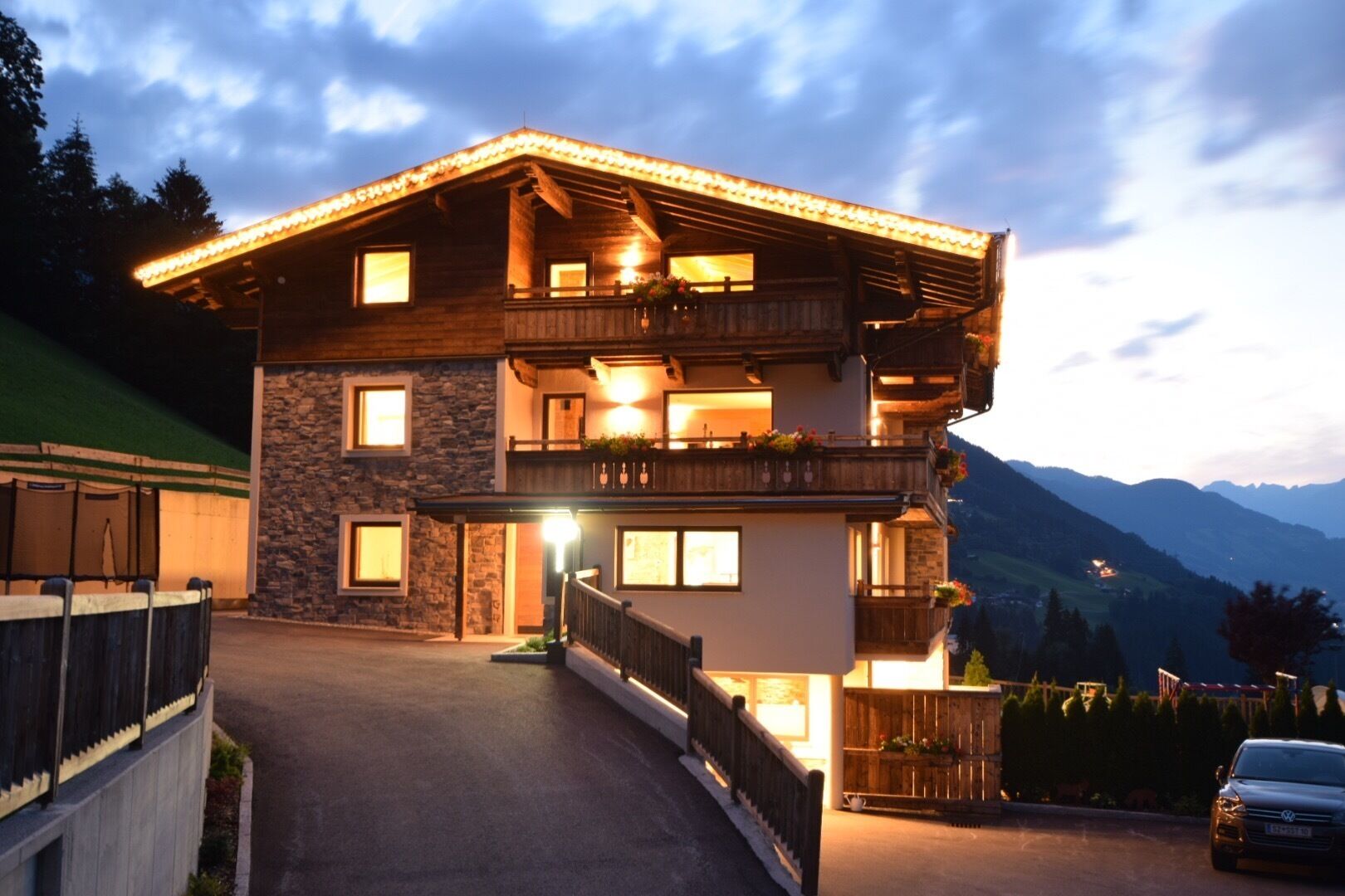 Un chalet à deux étages avec une façade en pierre, des balcons en bois et une allée menant à l’entrée.