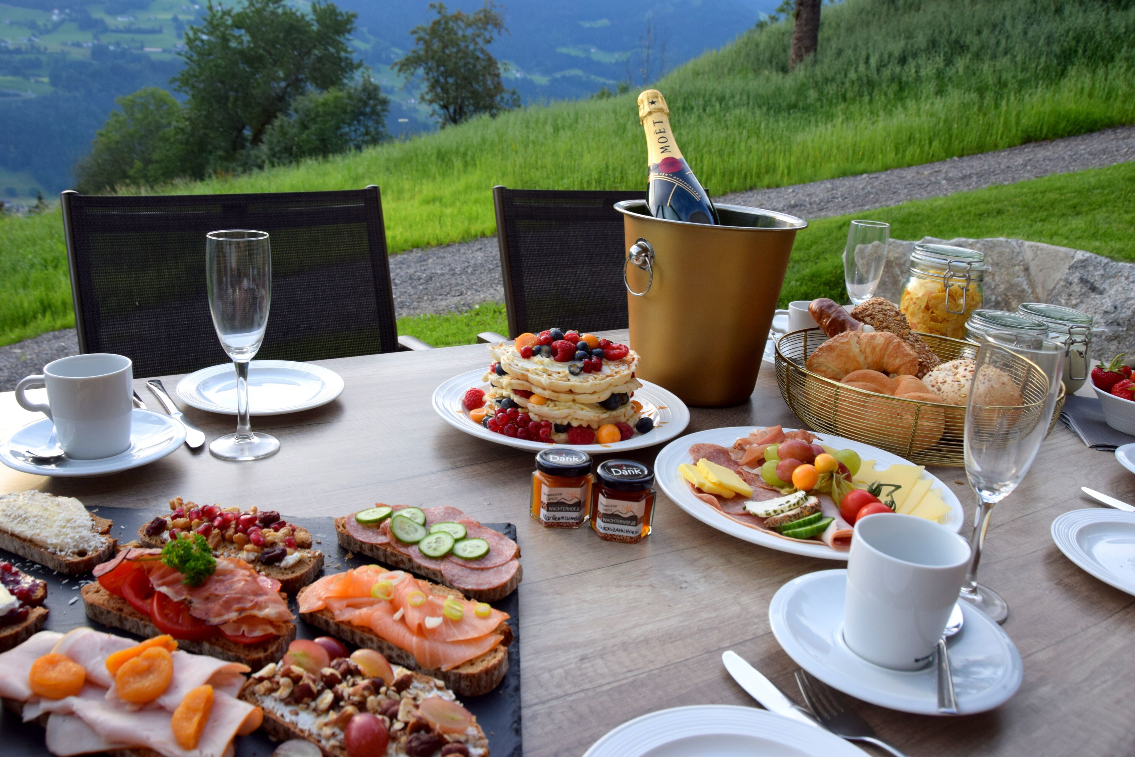 A table set for breakfast with various foods, a bottle of champagne, and a scenic view.