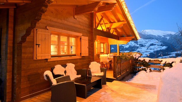 Une cabane en bois dotée d’un porche couvert, aménagée avec des chaises et une table, et offrant une vue sur un paysage montagneux enneigé.