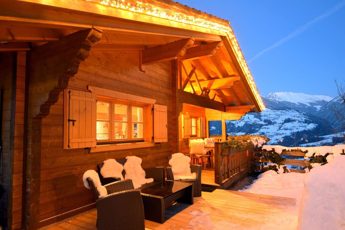 A wooden cabin with a covered porch, furnished with chairs and a table, overlooking a snowy mountain landscape.