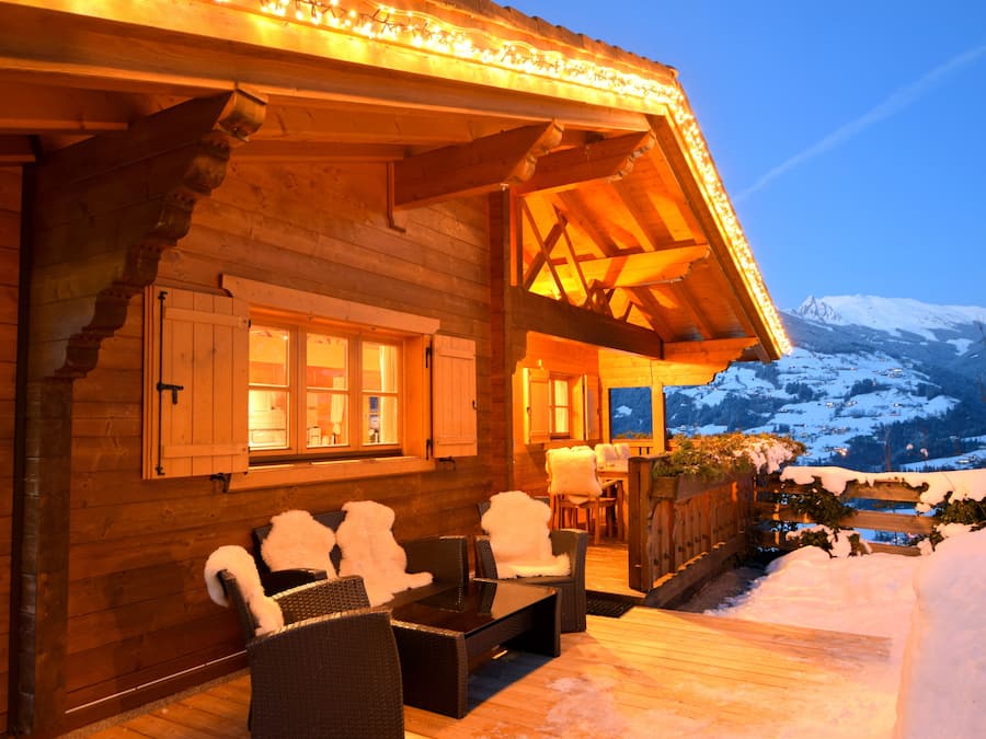 Una cabaña de madera con un porche cubierto, amueblada con sillas y una mesa, con vistas a un paisaje montañoso nevado.