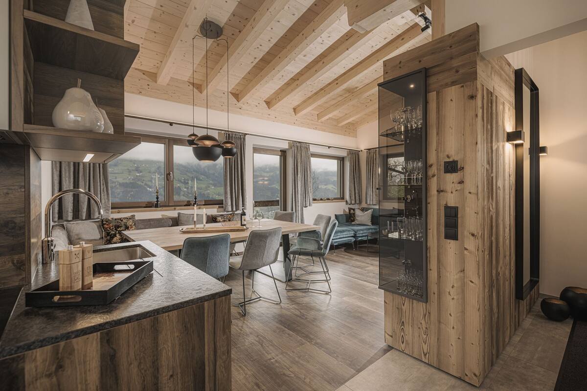 A modern kitchen with wooden beams, a dining area with chairs, and a view of the countryside through large windows.
