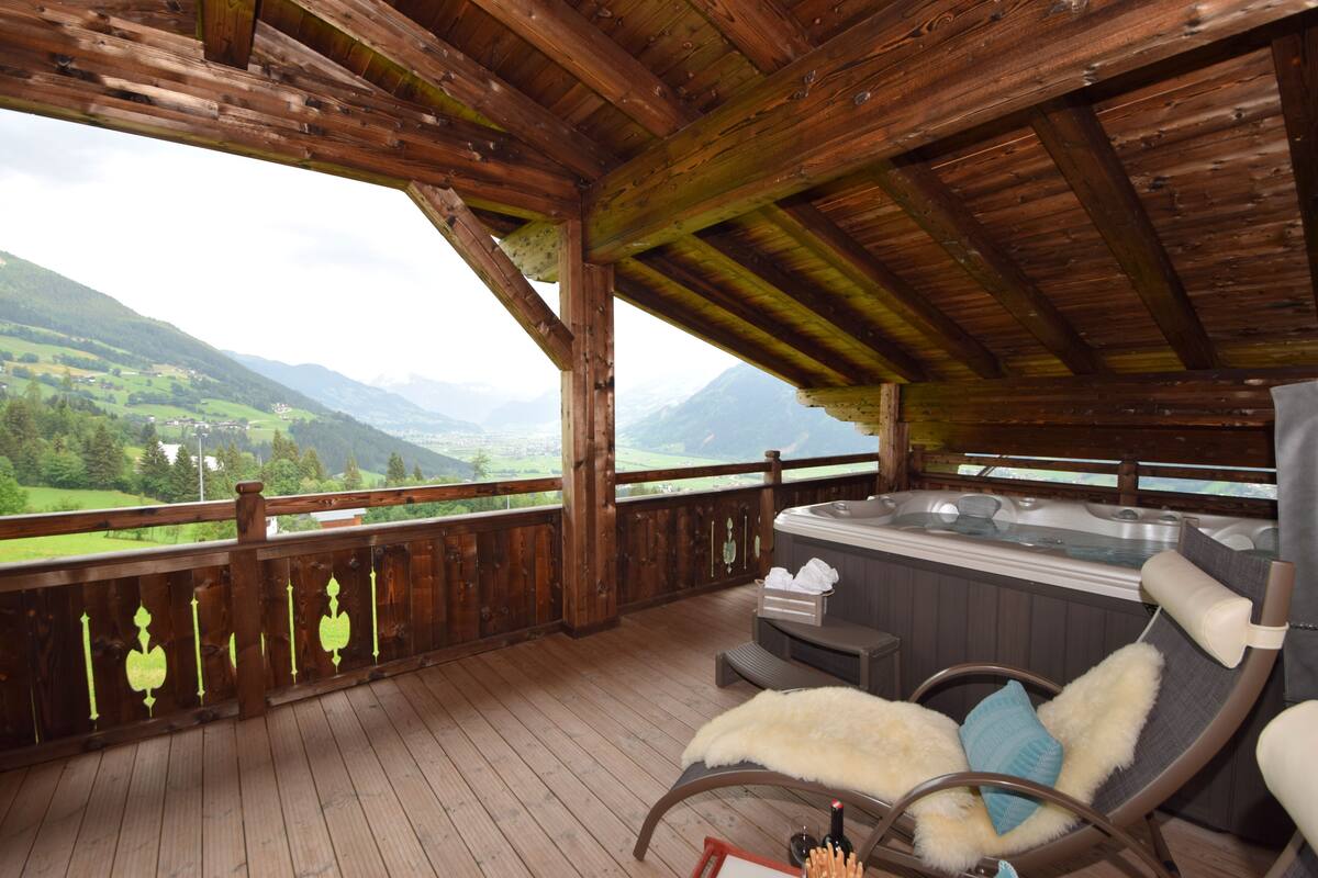 A wooden deck with a hot tub, a cushioned chair, and a scenic mountain view.