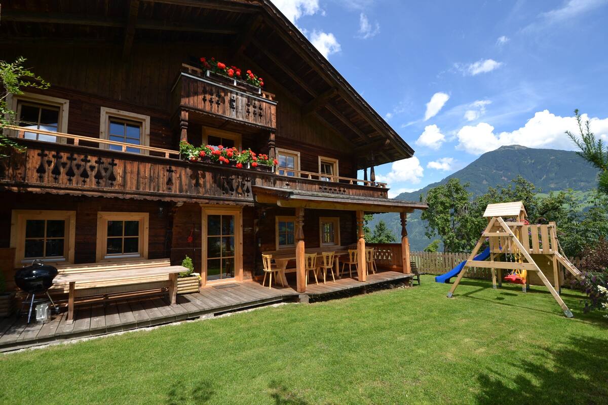 A wooden chalet with a balcony, a barbecue area, and a playground.