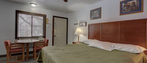 A hotel room with a bed, a desk with a chair, a ceiling fan, and a window with blinds.