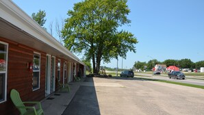 Ein Motel mit einer Reihe von Zimmern, einem überdachten Außenbereich zum Sitzen und einem großen Baum davor.