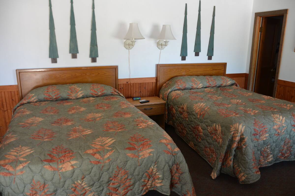 A hotel room with two single beds, wooden headboards, a nightstand with a lamp, and a door to the right.