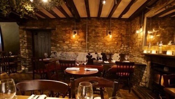Un restaurant chaleureux aux murs de pierre, aux poutres apparentes et aux luminaires suspendus. Les tables sont dressées avec des verres à vin et des couverts.
