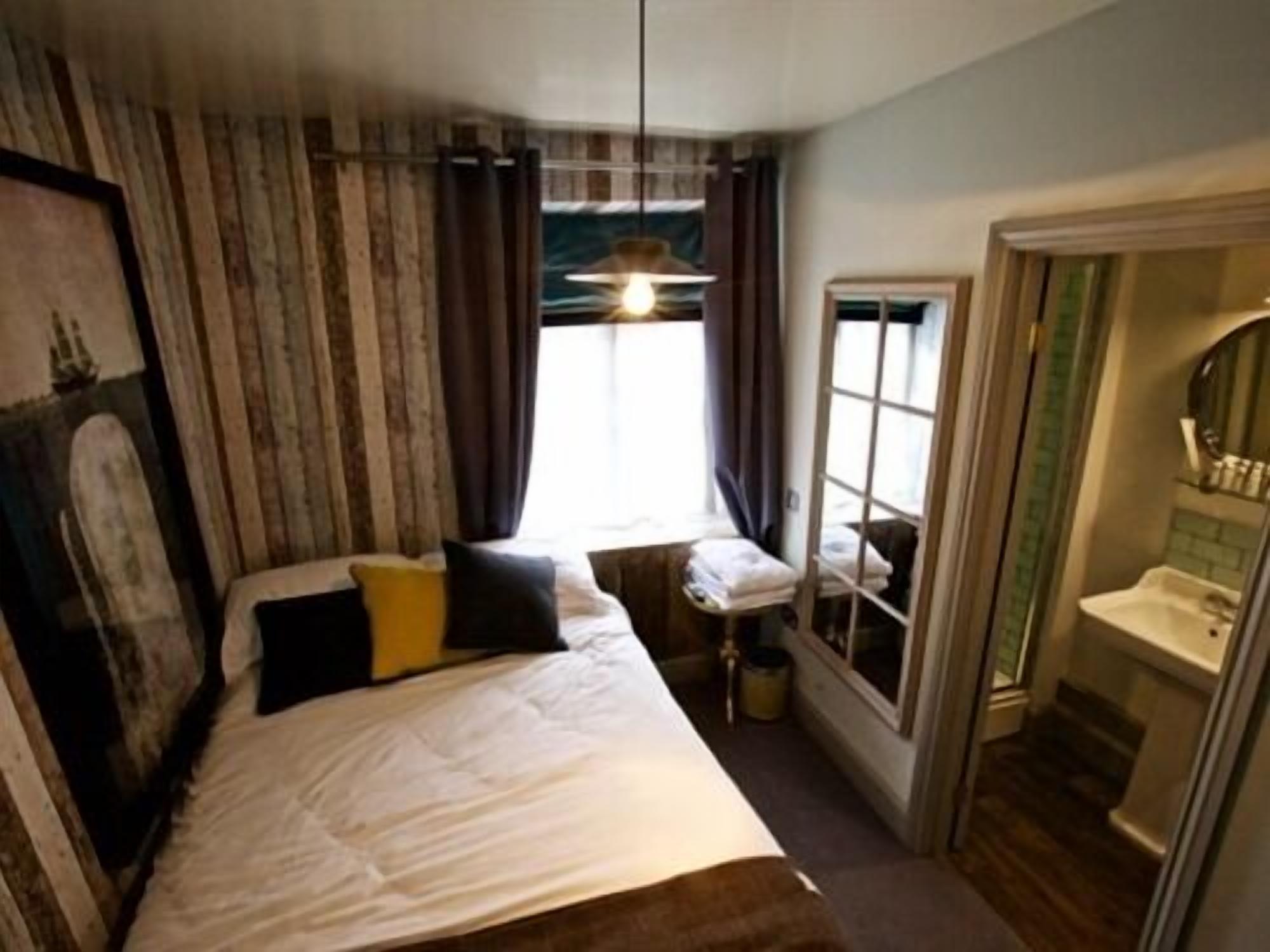 A bedroom with a bed, a window with curtains, a bedside table, and a bathroom with a sink and mirror.