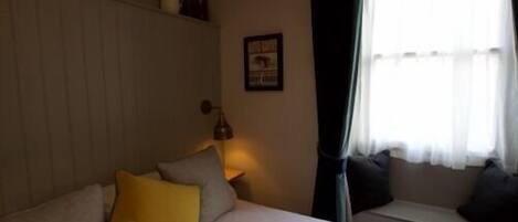 A bedroom with a bed, pillows, a nightstand, a lamp, a window with curtains, and a wall-mounted picture.