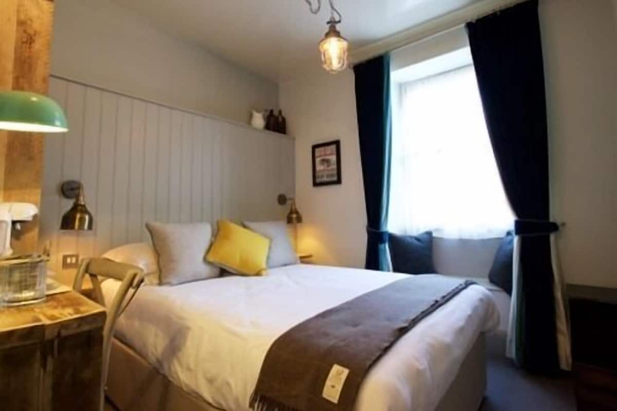 Un dormitorio ordenado con cama, mesita de noche, lámpara y ventana con cortinas.