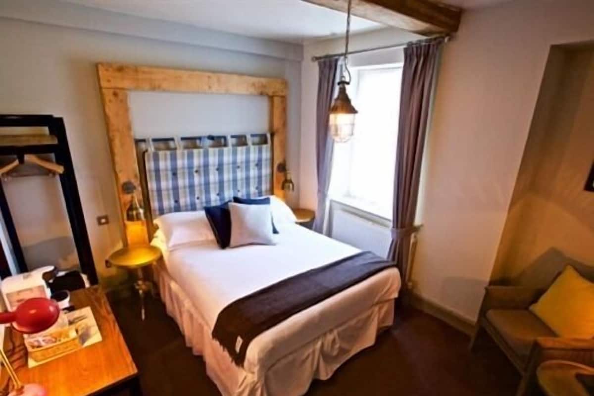 Habitación de hotel ordenada con cama, escritorio con lámpara, silla y ventana con cortinas.