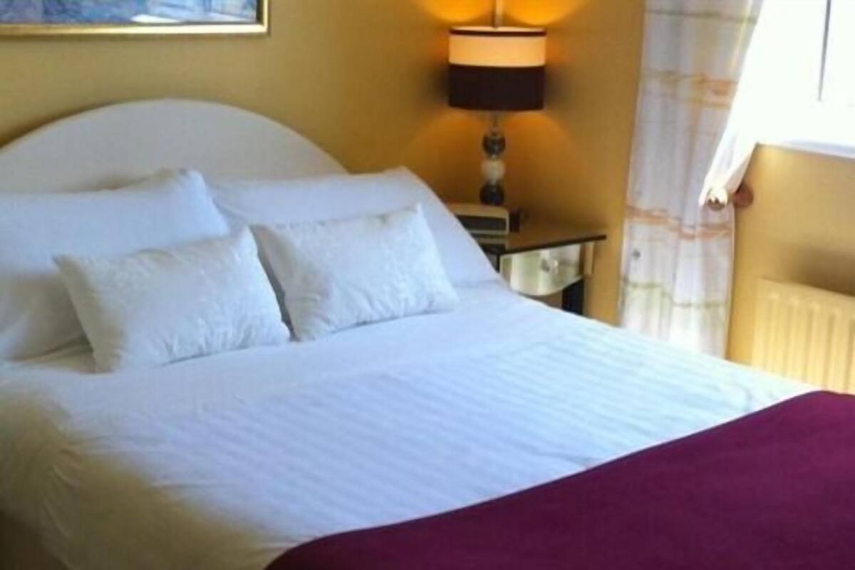 Uma cama com roupa de cama branca e um cobertor vermelho, uma luminária de cabeceira e uma janela com cortinas.