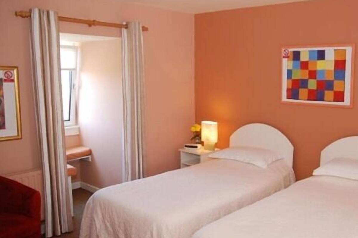 Quarto de hotel com duas camas, uma mesa de cabeceira com abajur, uma obra de arte colorida na parede e uma cadeira vermelha.