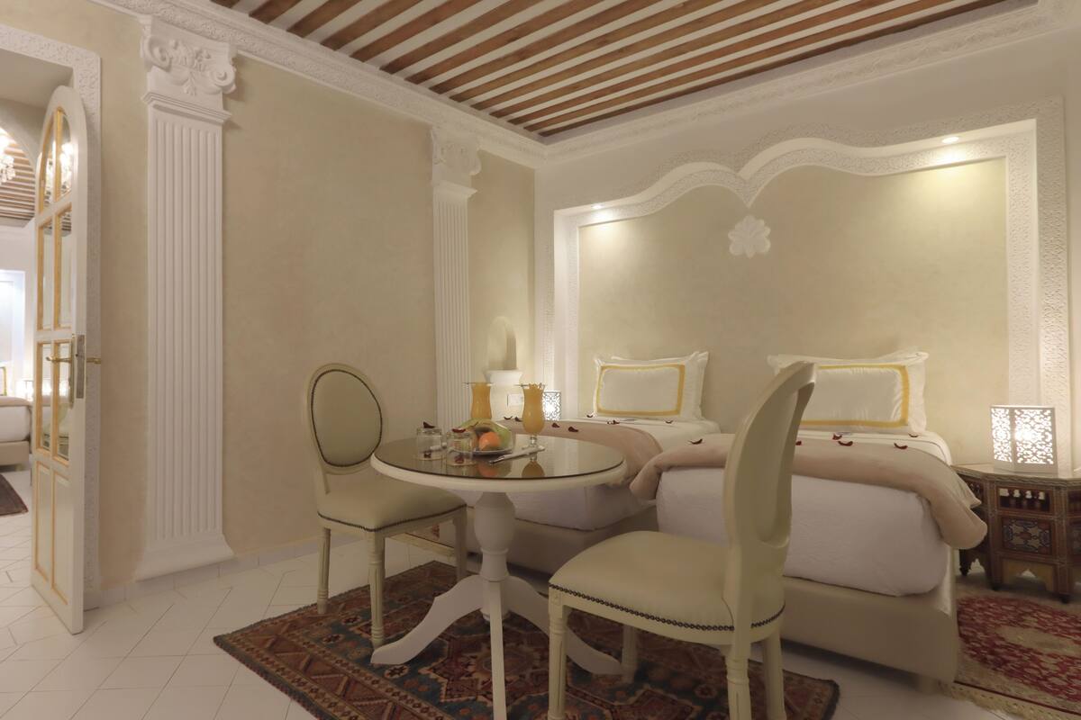 Habitación de hotel con cama, una mesa redonda con sillas, una mesita de noche con lámpara y un espejo decorativo.
