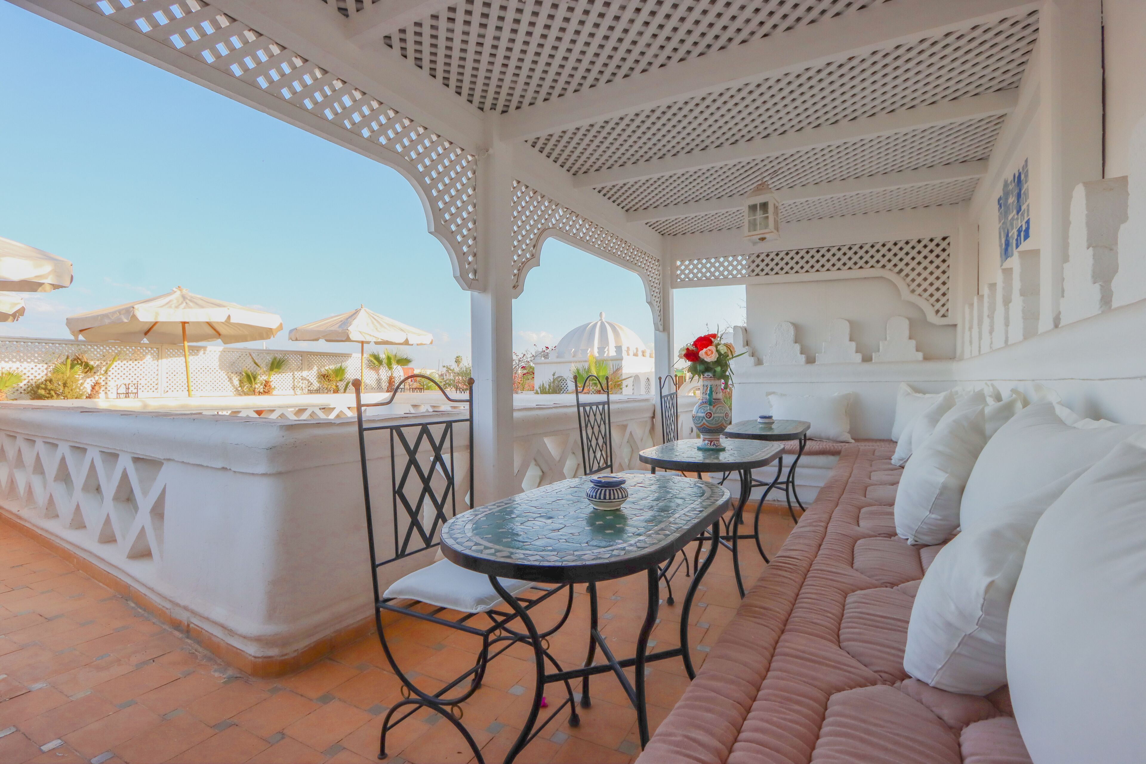 A covered outdoor seating area with tables and chairs, a white wall with a decorative pattern, and a view of a dome structure in the background.