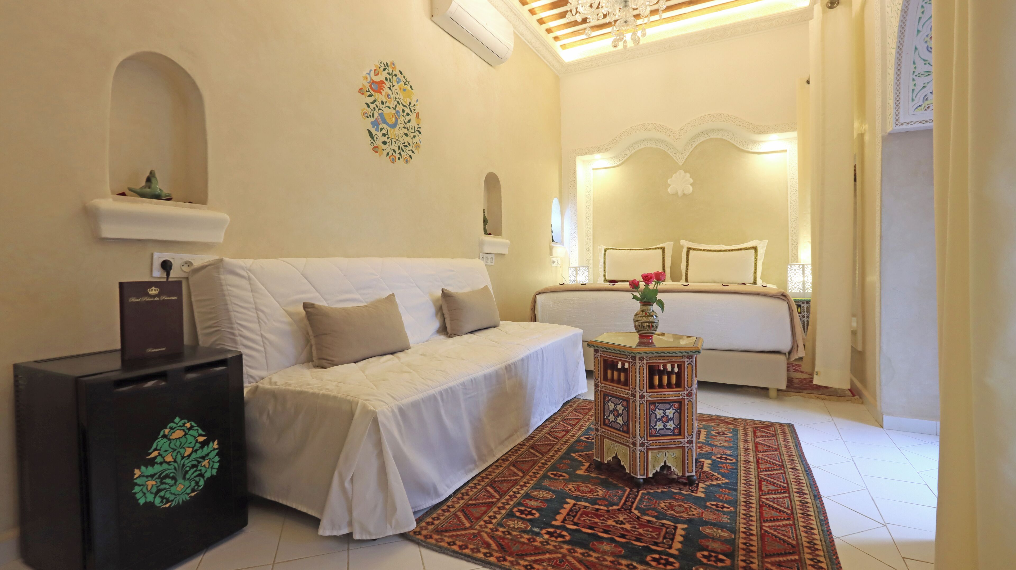 Habitación de hotel con un sofá blanco, una mesita con un jarrón, un tapete estampado y una cama con cabecero.