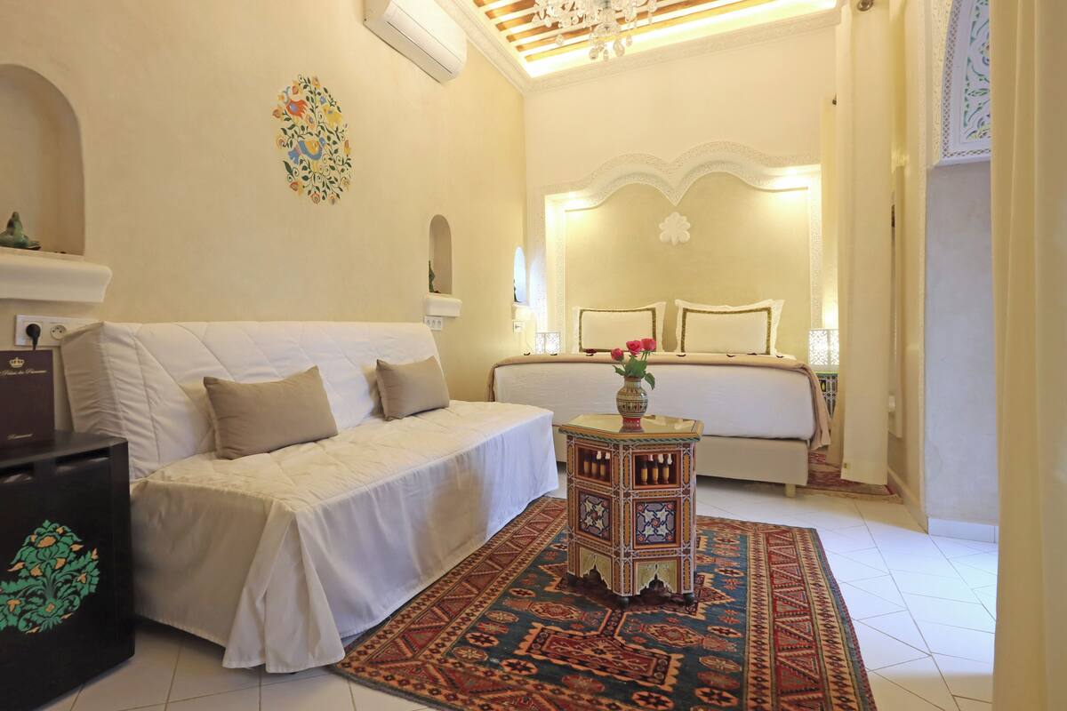 Habitación de hotel con un sofá blanco, una mesita con un jarrón, un tapete estampado y una cama con cabecero.