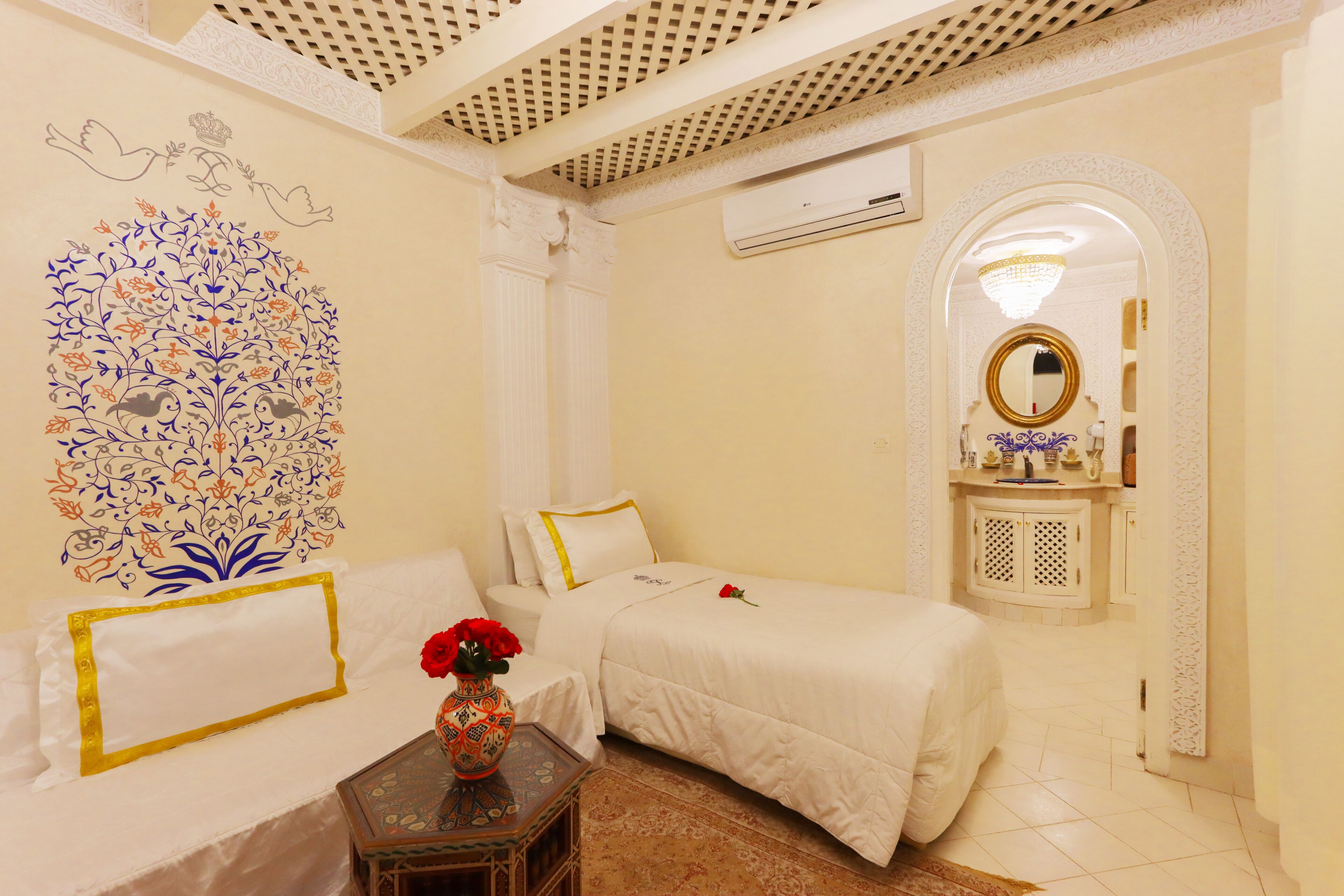 Quarto de hotel com duas camas, um mural decorativo, uma pequena mesa com um vaso, e banheiro com espelho e pia.