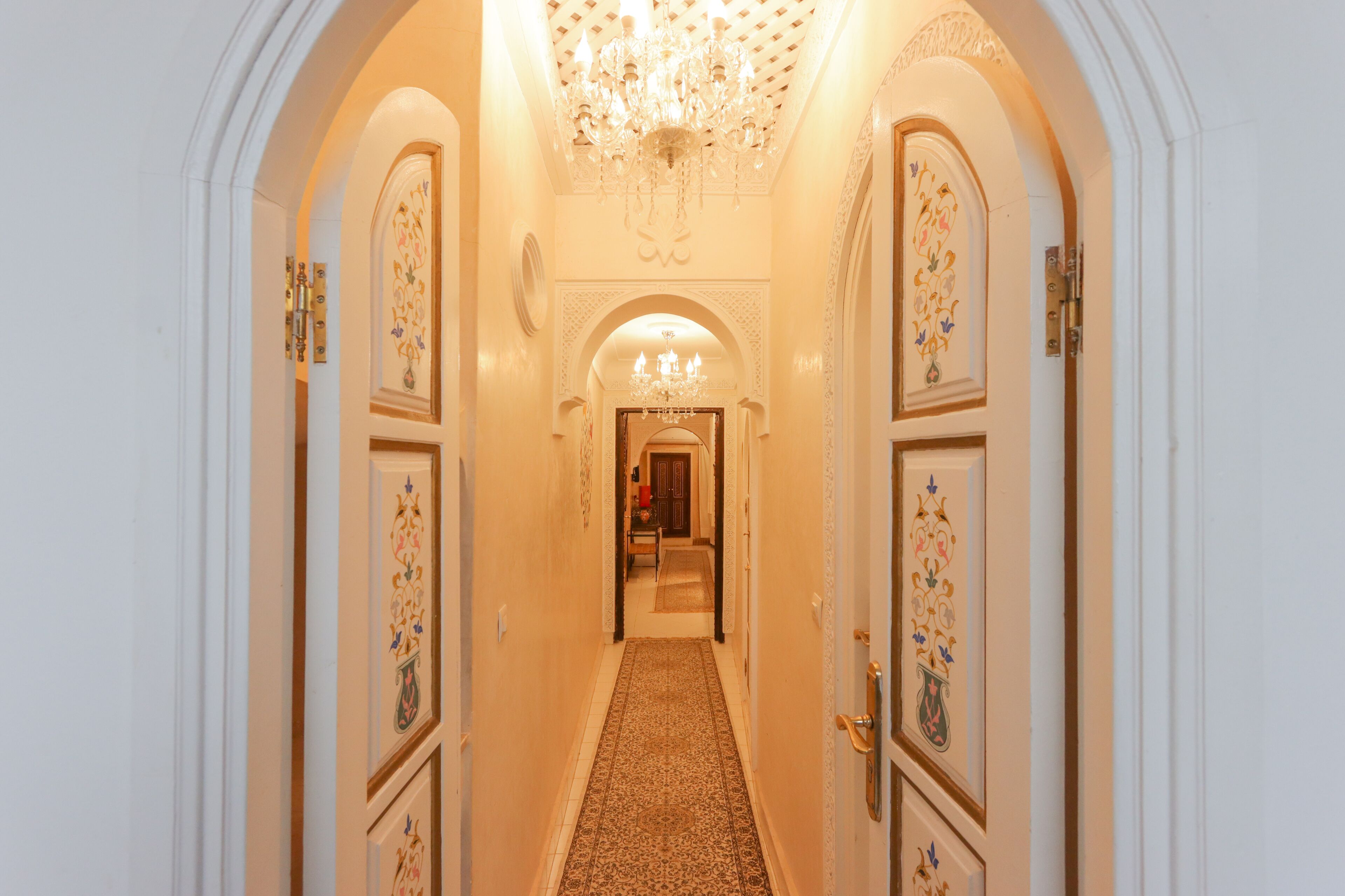 Un couloir orné de portes richement décorées, recouvert d’un tapis à motifs et illuminé par un lustre.