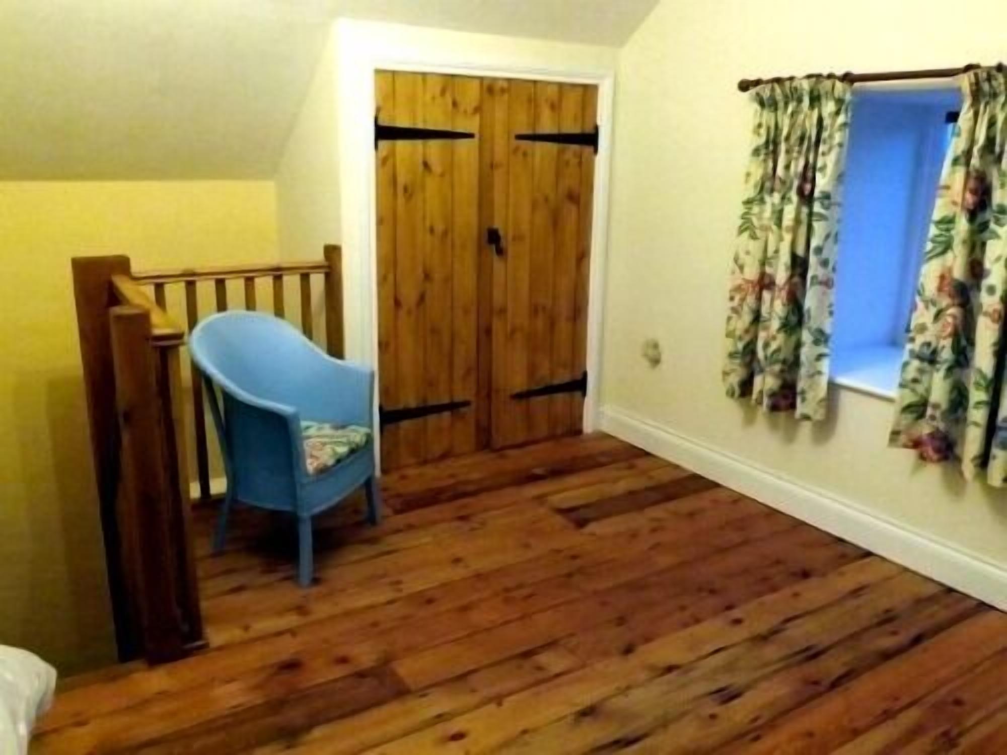 Una habitación con piso de madera, una silla azul, una puerta de madera y cortinas con motivos florales.
