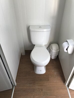 A small, white toilet with a lid, a white trash bin, and a toilet paper holder in a narrow bathroom with wooden flooring. - Bamboo Paracas Resort (Paracas)