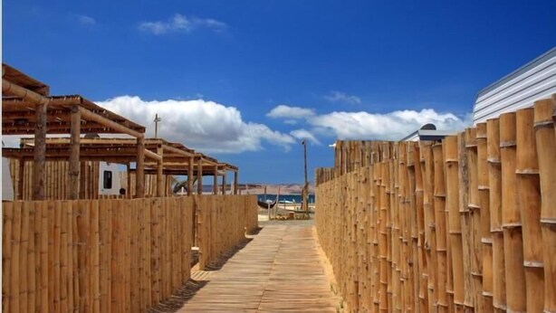 A wooden boardwalk leading through a rustic area with bamboo walls and a clear blue sky. - Bamboo Paracas Resort (Paracas)