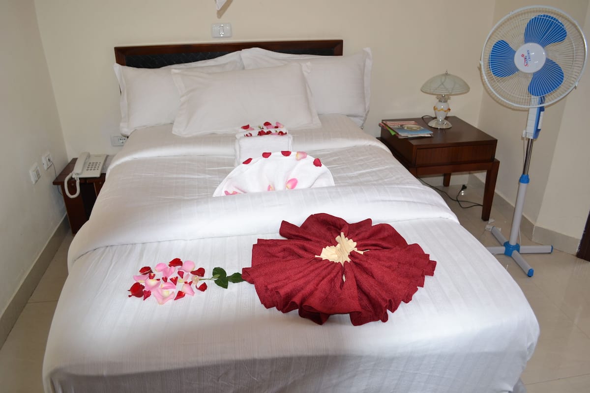 A neatly made bed with white linens, a red floral arrangement, and a bedside table with a lamp and a book.