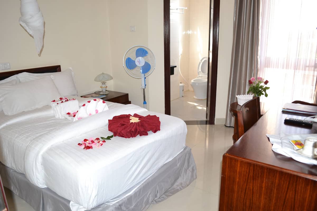 A hotel room with a bed, bedside table, lamp, fan, and a desk with a remote control.