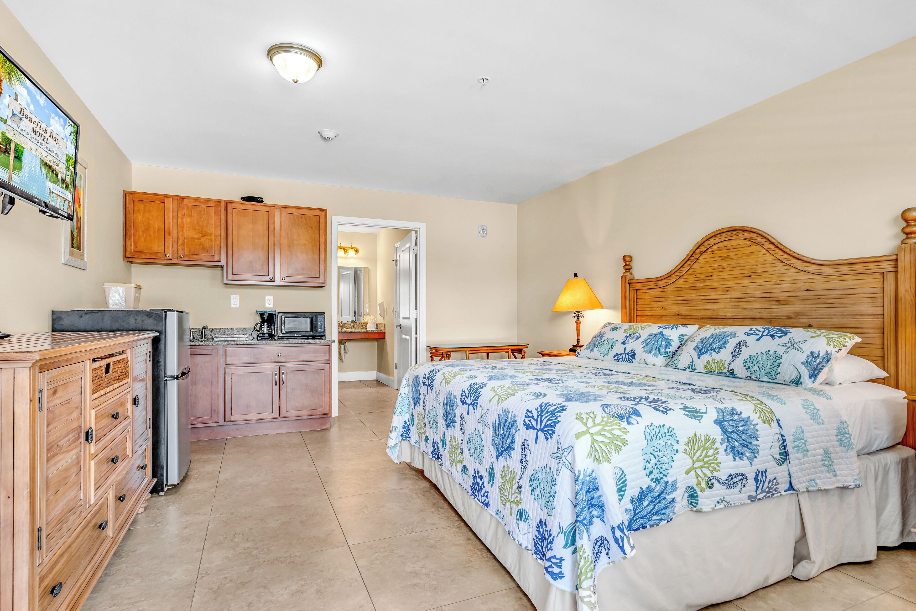 A bedroom with a bed, wooden headboard, and a nightstand. There is a kitchen area with cabinets and a refrigerator.