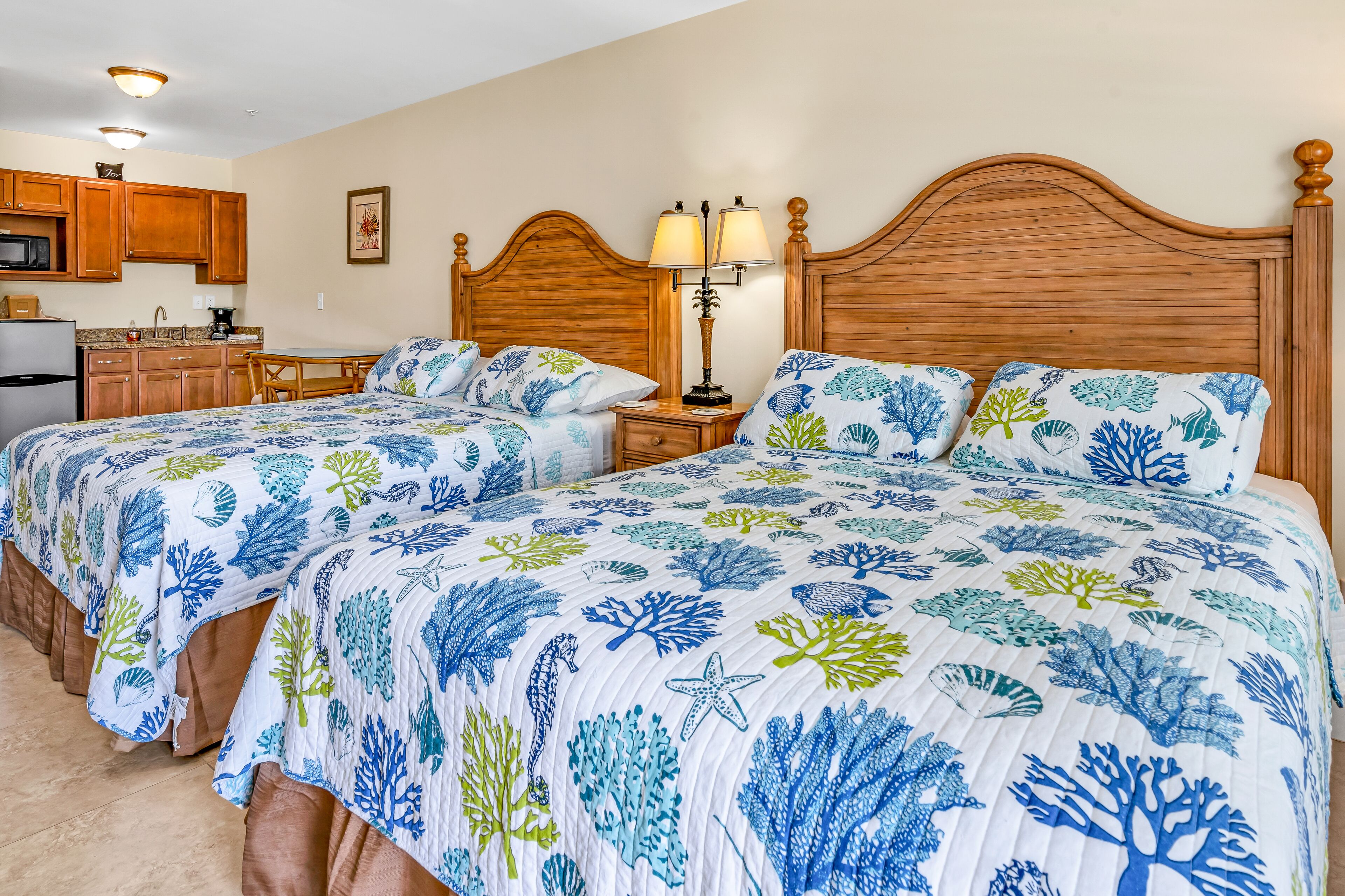 A bedroom with two beds, a wooden headboard, a nightstand with a lamp, a small kitchen area, and a microwave.