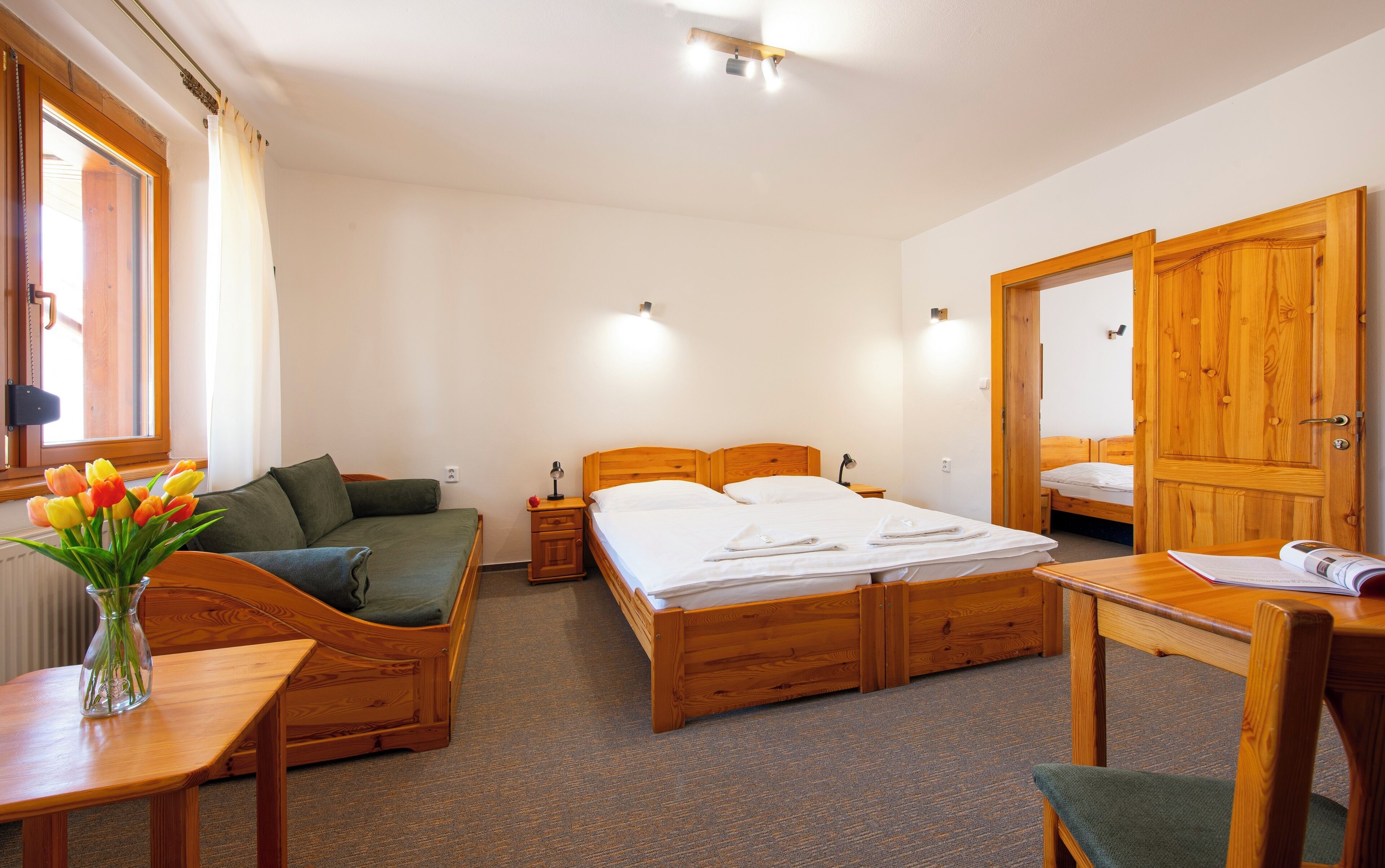 A hotel room with a wooden bed, a desk, a chair, a small table, a vase with flowers, and a window with curtains.